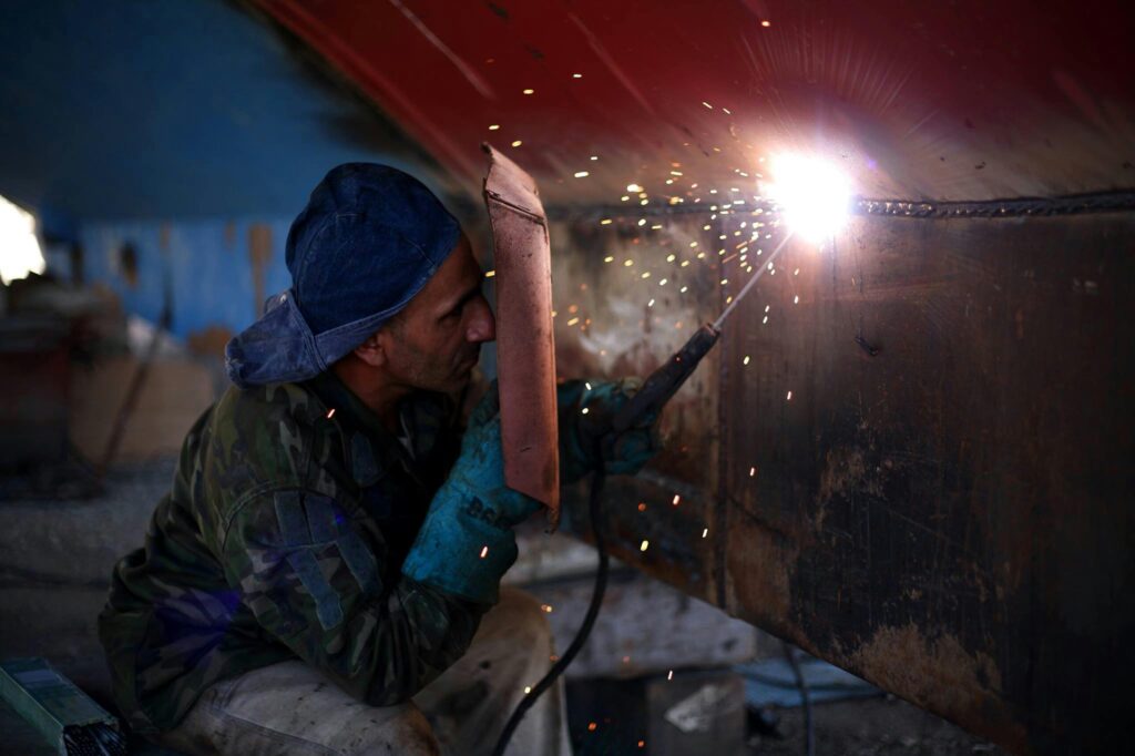 A welder in protective gear works indoors, generating sparks with a welding torch.