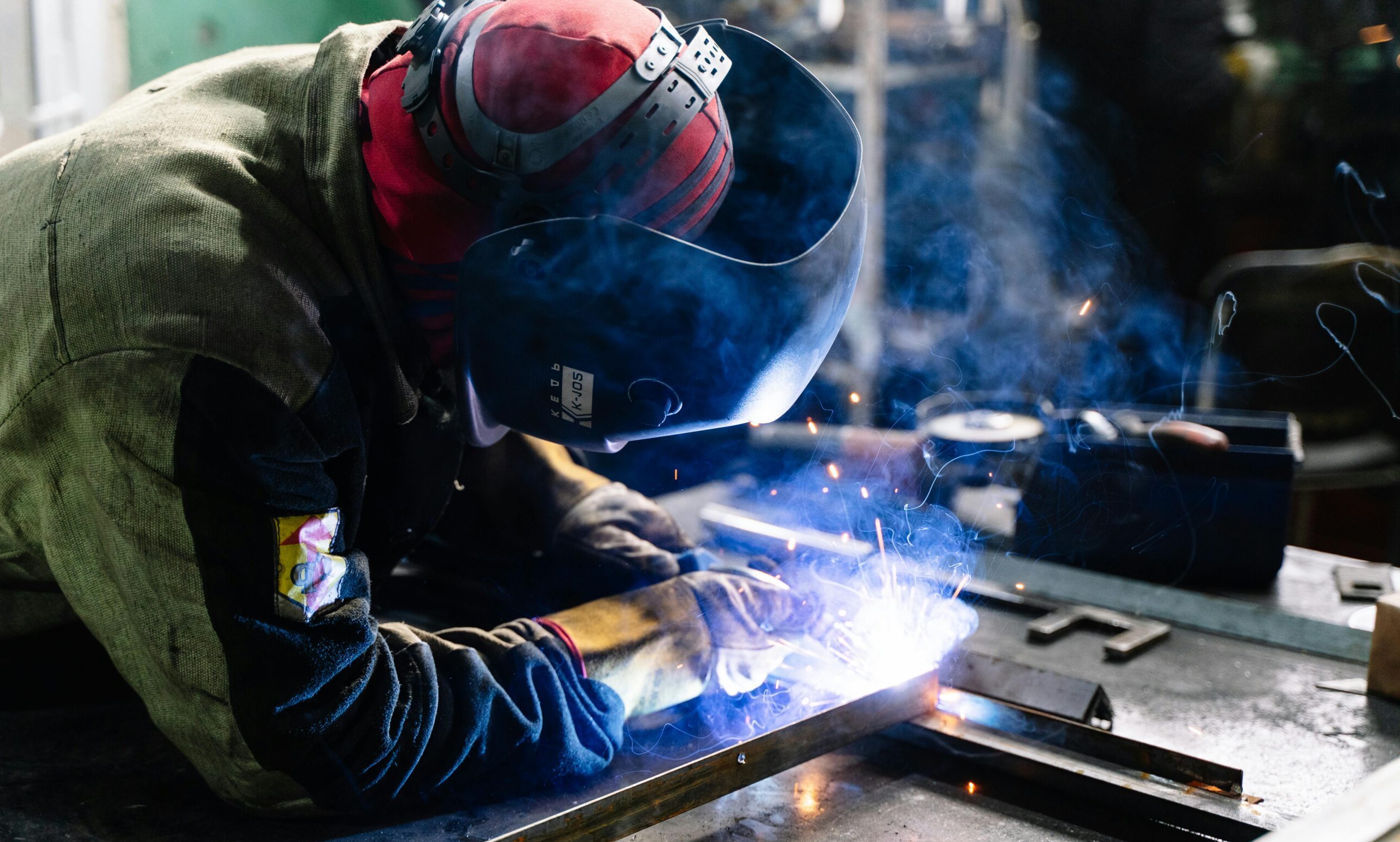 A welder works on metal fabrication indoors, creating bright sparks and smoke.